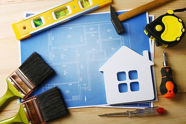 Construction tools, paintbrushes, and a white house model arranged on top of blue architectural blueprints.