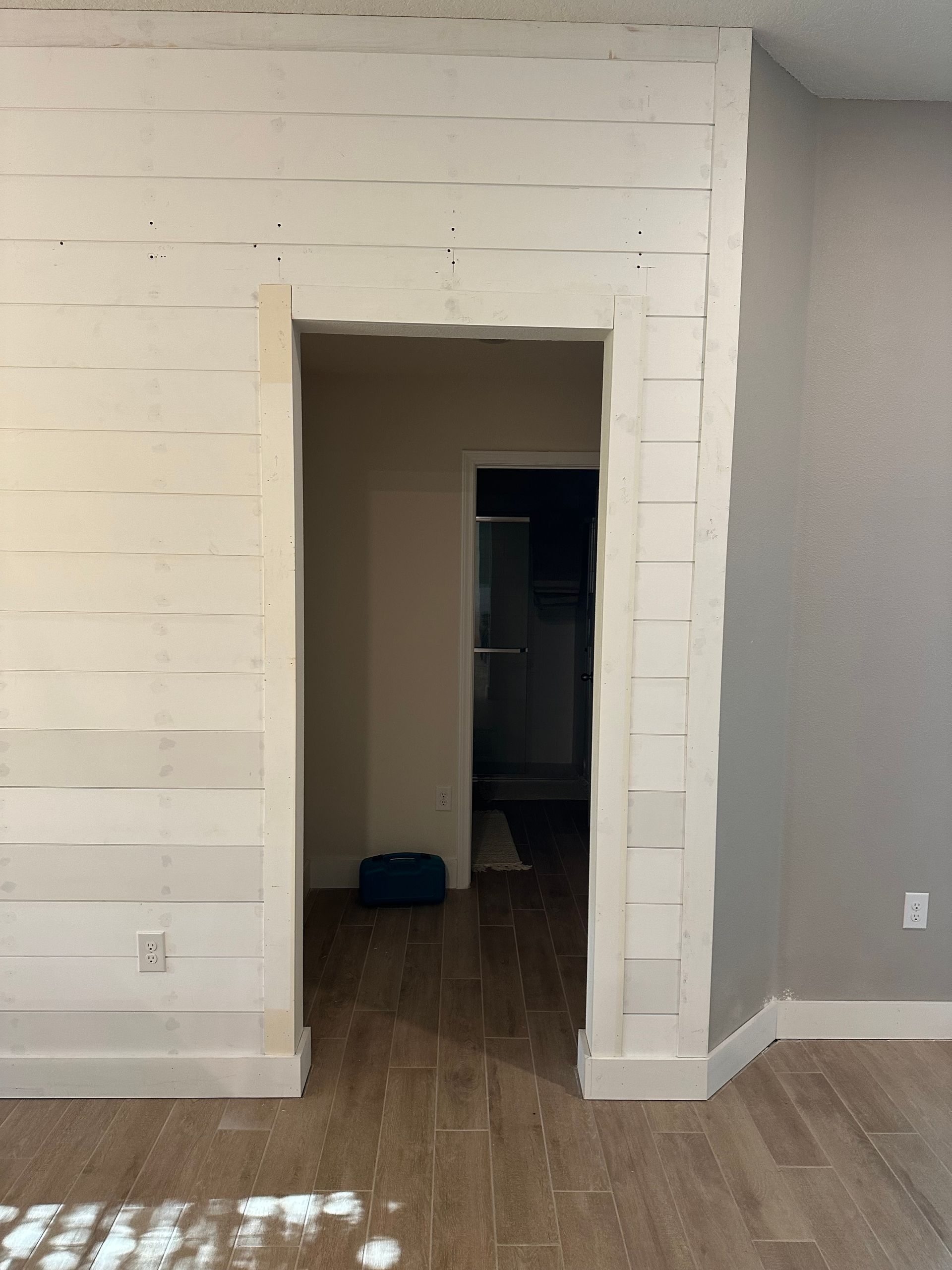 Interior doorway with white shiplap walls, leading to a room with dark doorway and grey wall.