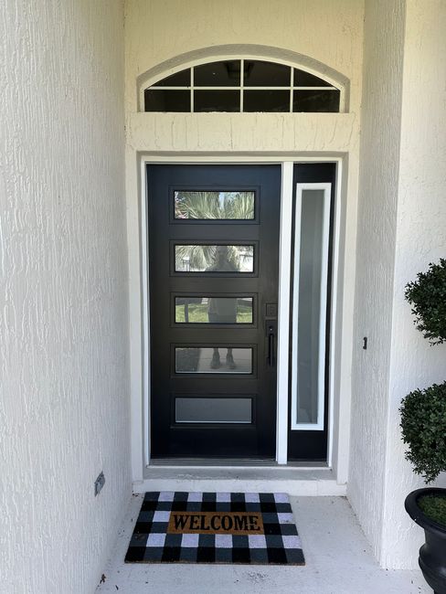 Black front door with glass panels, sidelight, and welcome mat; set in a stucco facade.