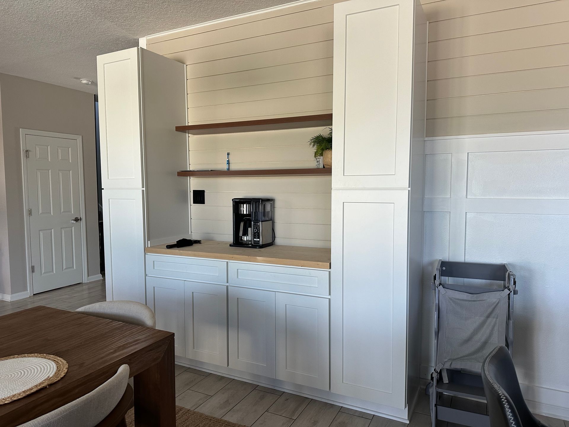 White built-in cabinets featuring a coffee station, open wooden shelving, and shiplap wall detail in a dining room.