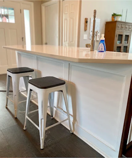 Two white stools sit at a white kitchen island with a light countertop. A doorway is visible behind.