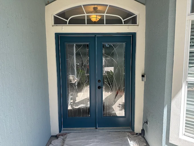 Blue double doors with decorative glass, beneath a matching arched transom, on a teal-colored house.