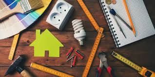 Home improvement tools, including a green house icon, lightbulb, tape measure, ruler, and paintbrush on a wooden surface.