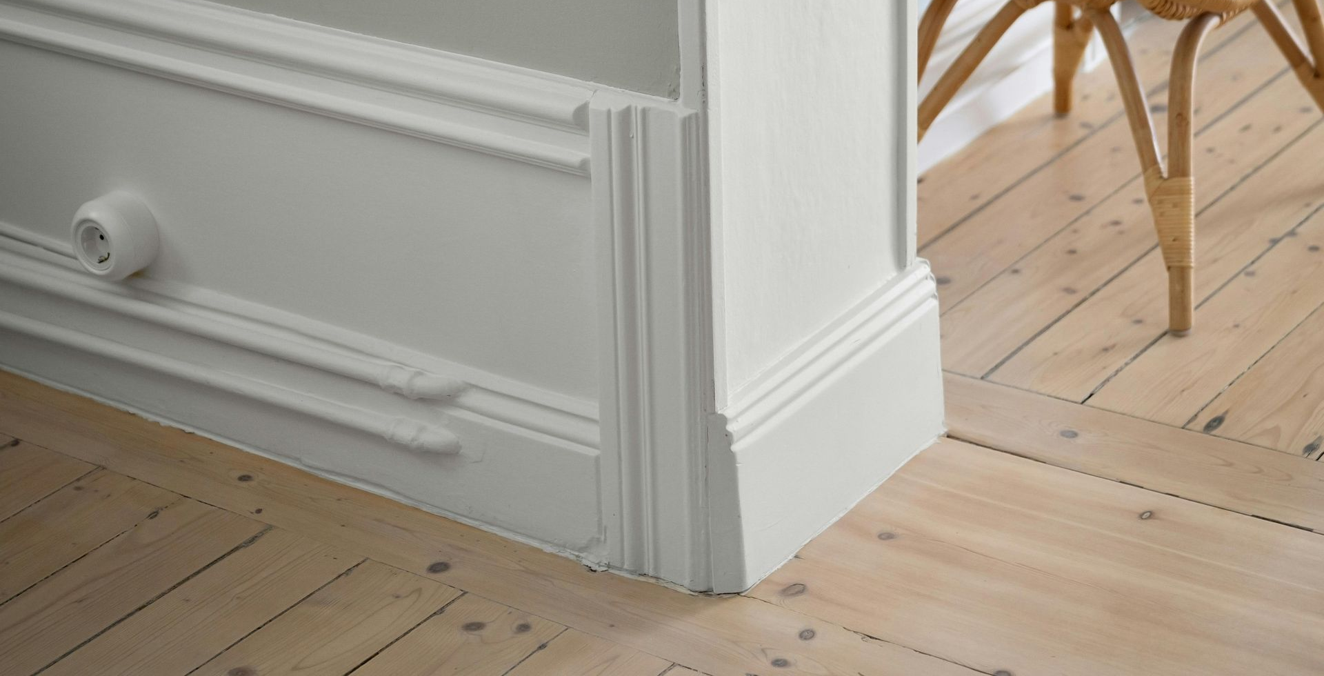 White wall with molding detail, corner of a room, and a woven stool on a wood floor.