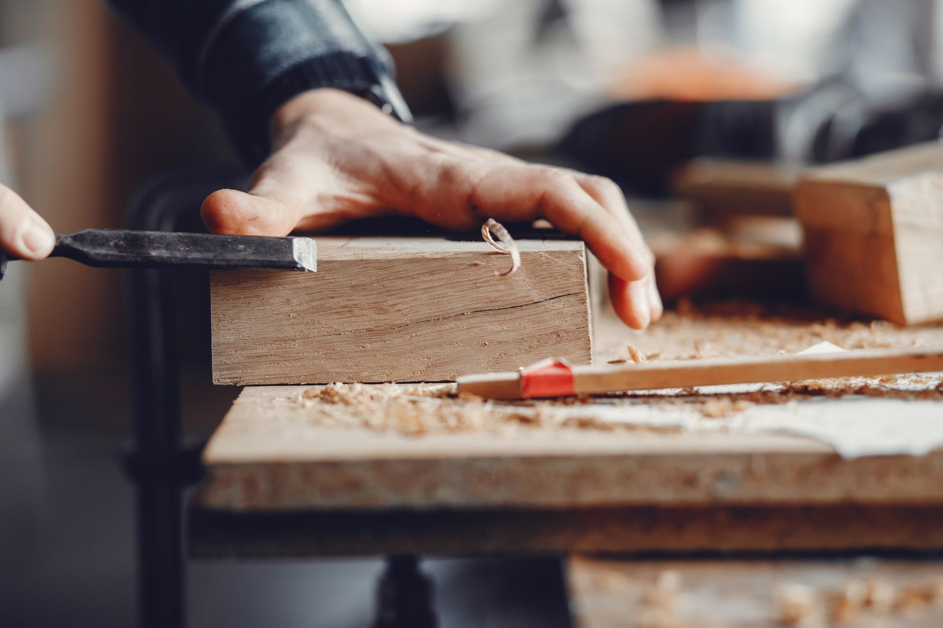 Person using a chisel to shape a wooden block on a workbench, with wood shavings.