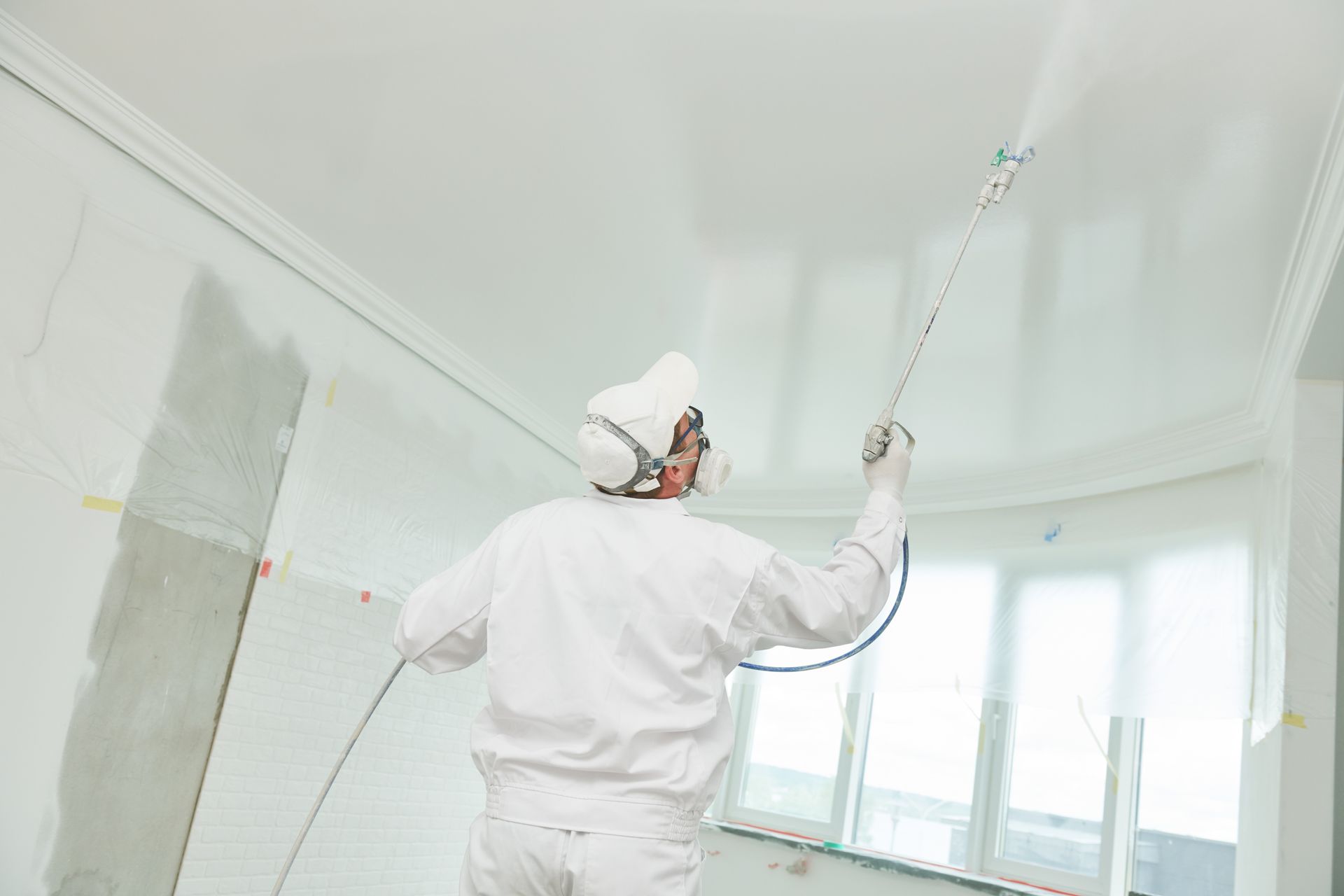 Person in protective suit sprays white paint on a ceiling with a paint sprayer.