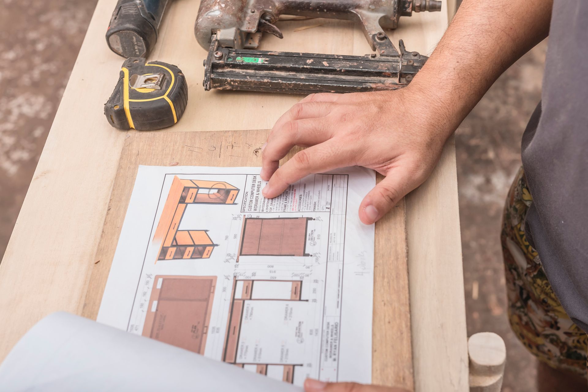 Person's hands on building plans with tools like a nail gun and measuring tape on a wooden surface.