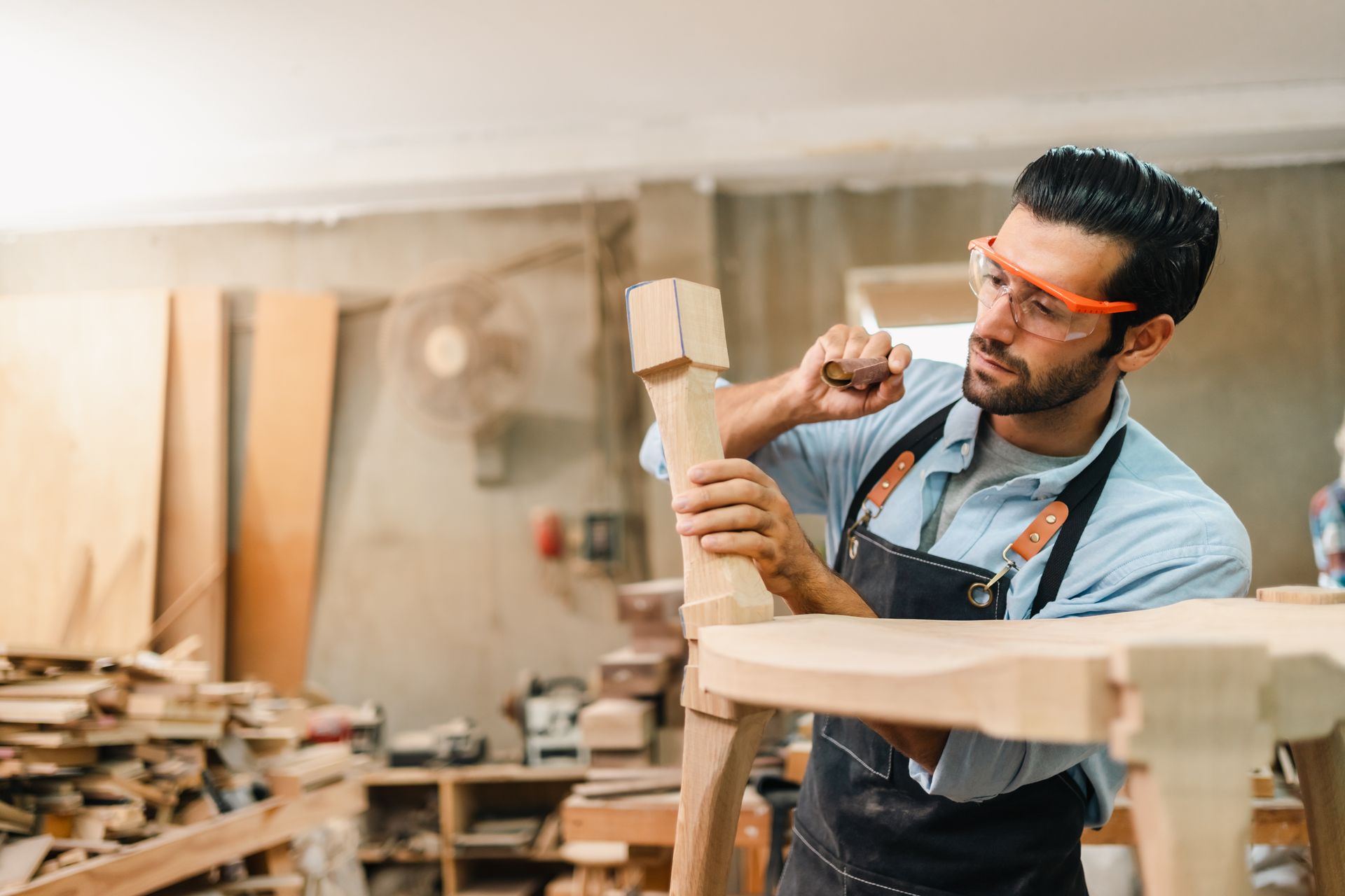 A person wearing safety glasses and an apron uses a chisel to shape a wooden furniture leg in a workshop.