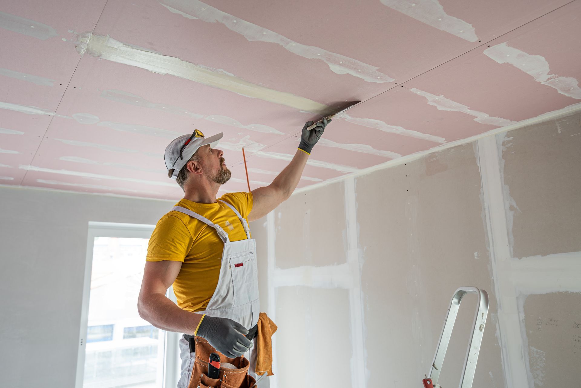 Man in yellow shirt and overalls, taping a drywall ceiling with a trowel, near a stepladder.