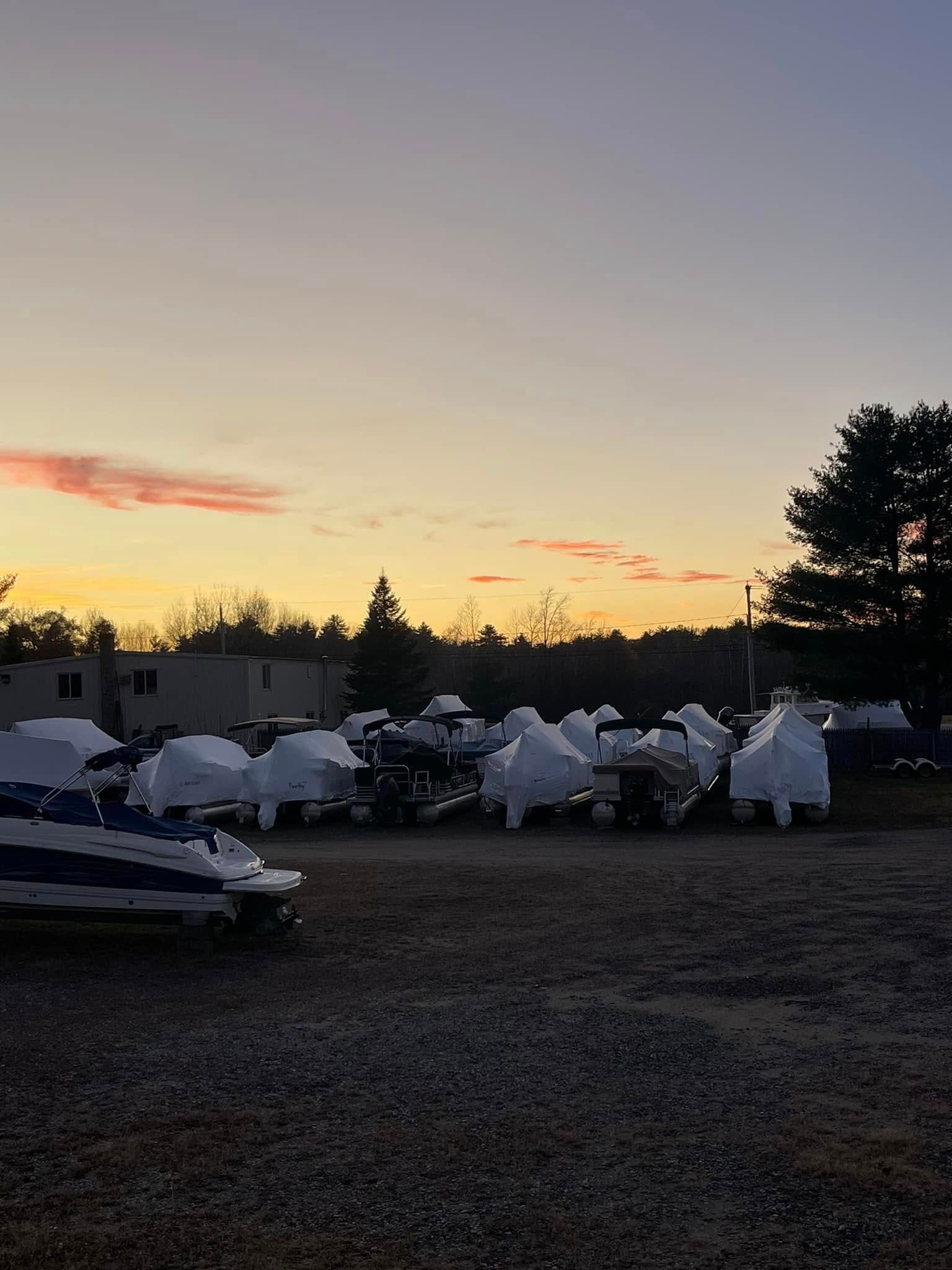 A lot of boats are parked in a gravel lot at sunset.
