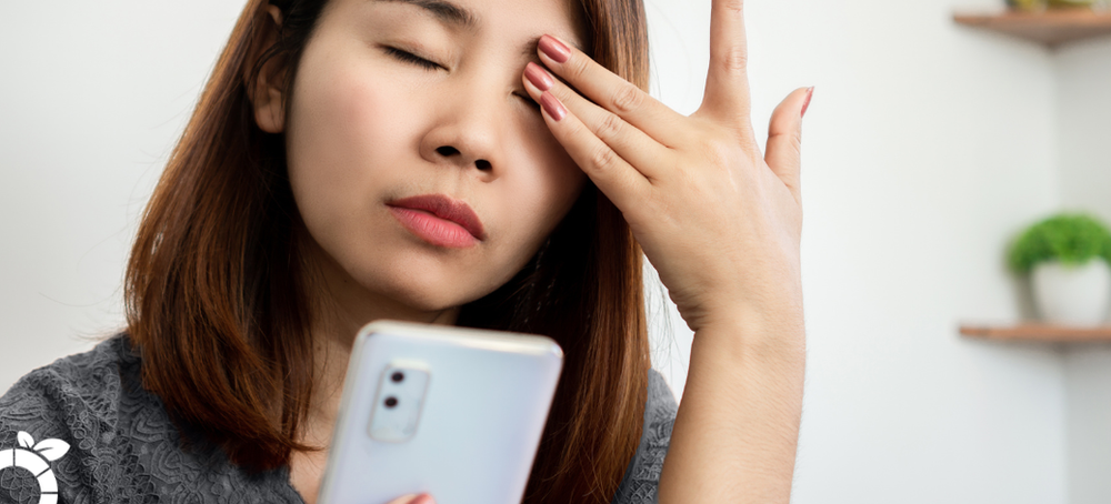 A woman is rubbing her eyes while looking at her cell phone.