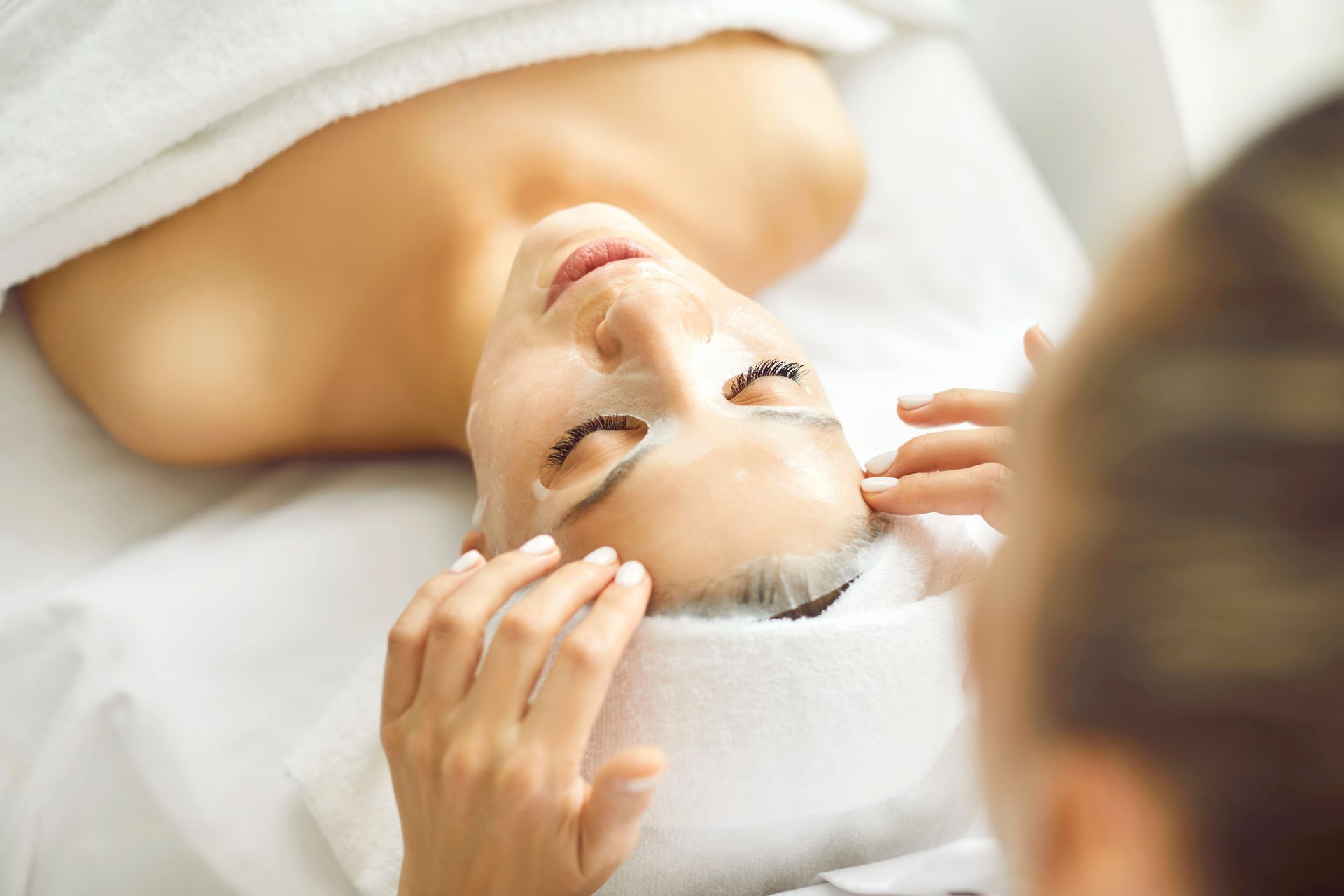 Woman receiving facial treatment; eyes closed, hands gently touching face, white towel, spa setting.