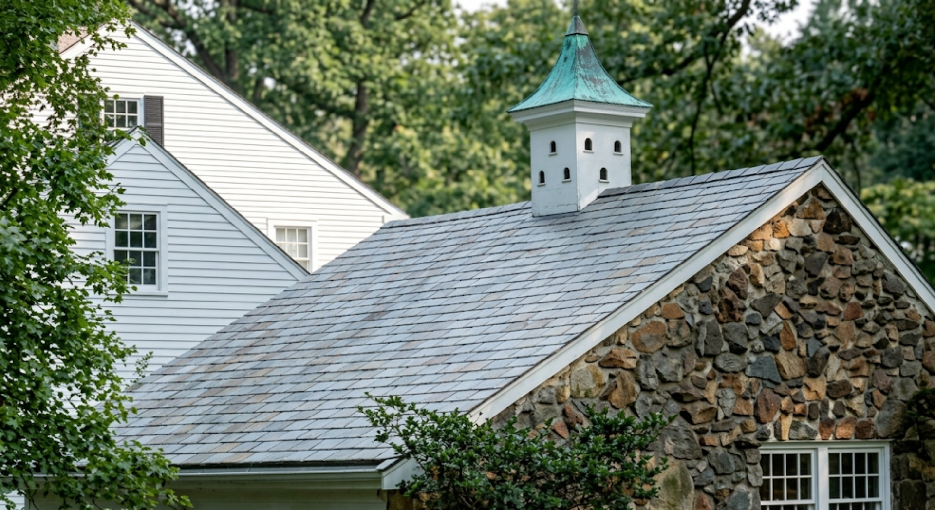 Stone house roof with gray shingles and a small green-topped cupola, framed by trees.