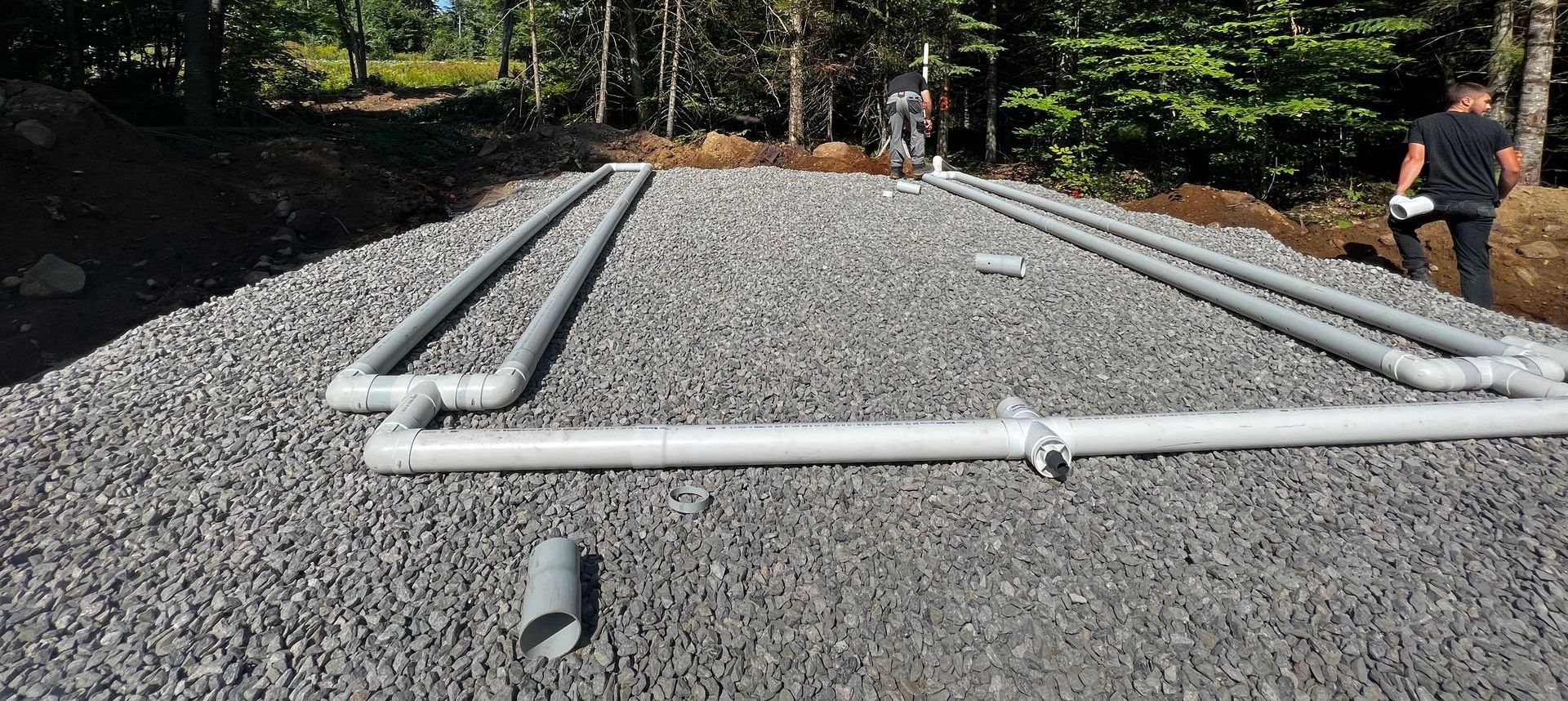 Lit de gravier avec des tuyaux gris, probablement pour l'installation d'une fosse septique, à l'extérieur. Une personne se tient à proximité.