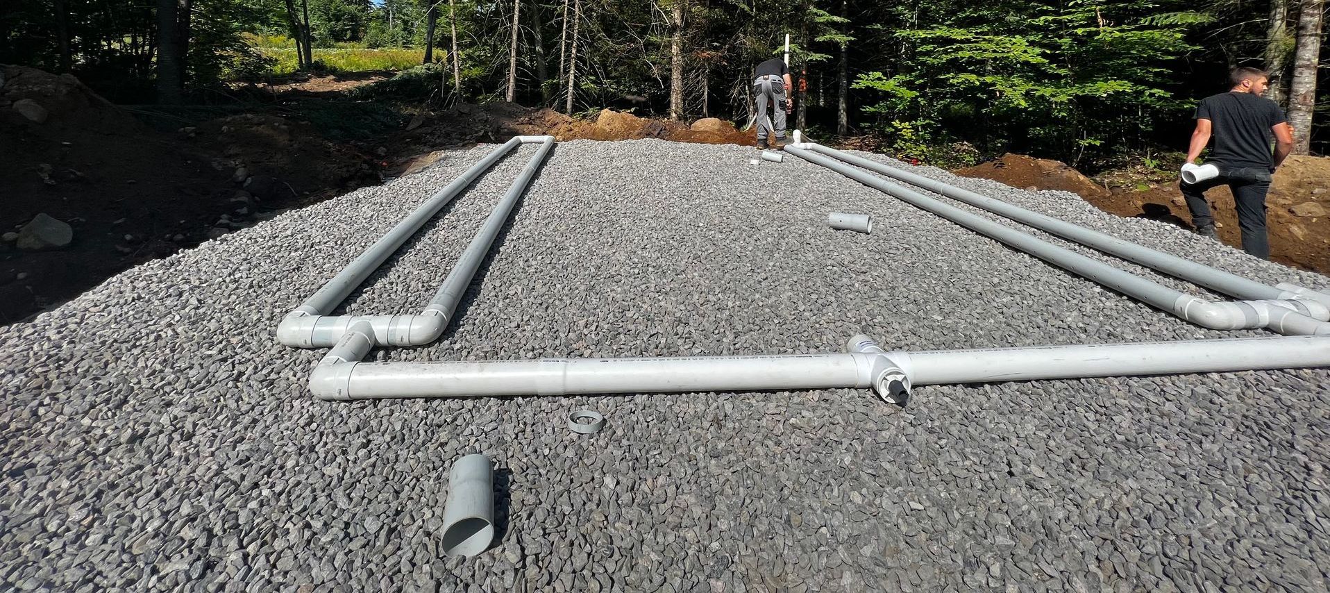 Lit de gravier avec tuyaux en PVC installés, probablement pour le drainage, dans un cadre extérieur arboré. Des personnes sont visibles en arrière-plan.