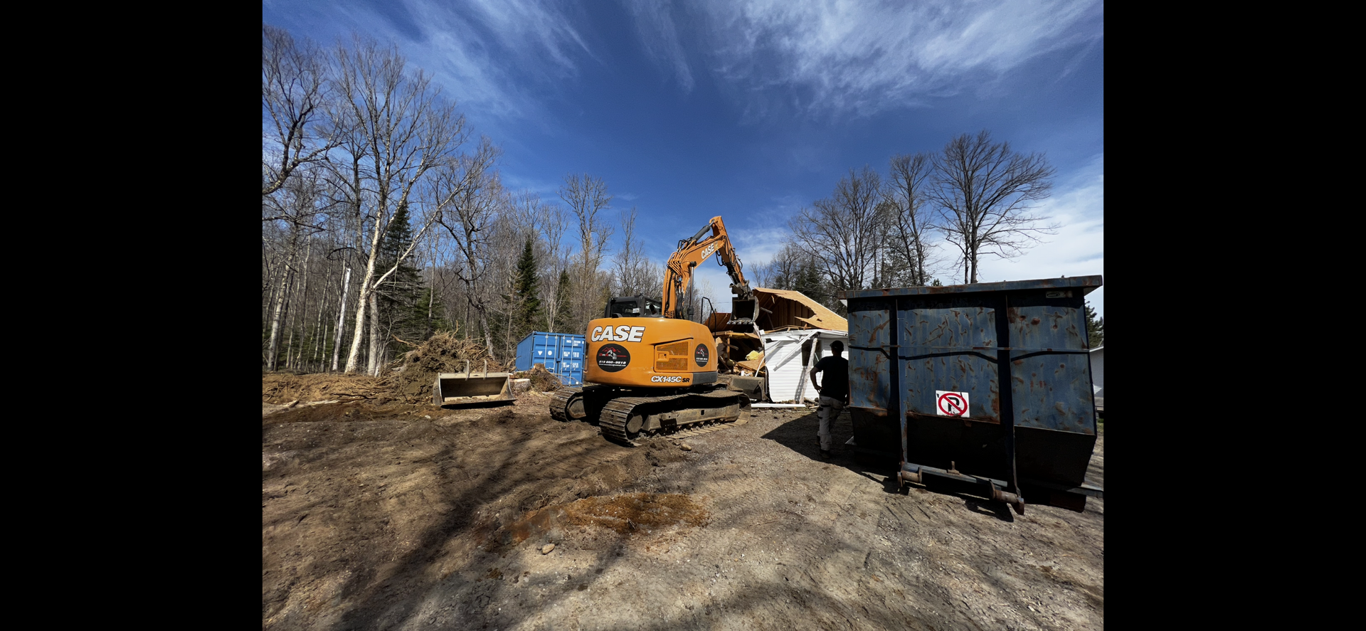 Une grande excavatrice jaune se trouve dans un champ de terre à côté d’une benne à ordures.
