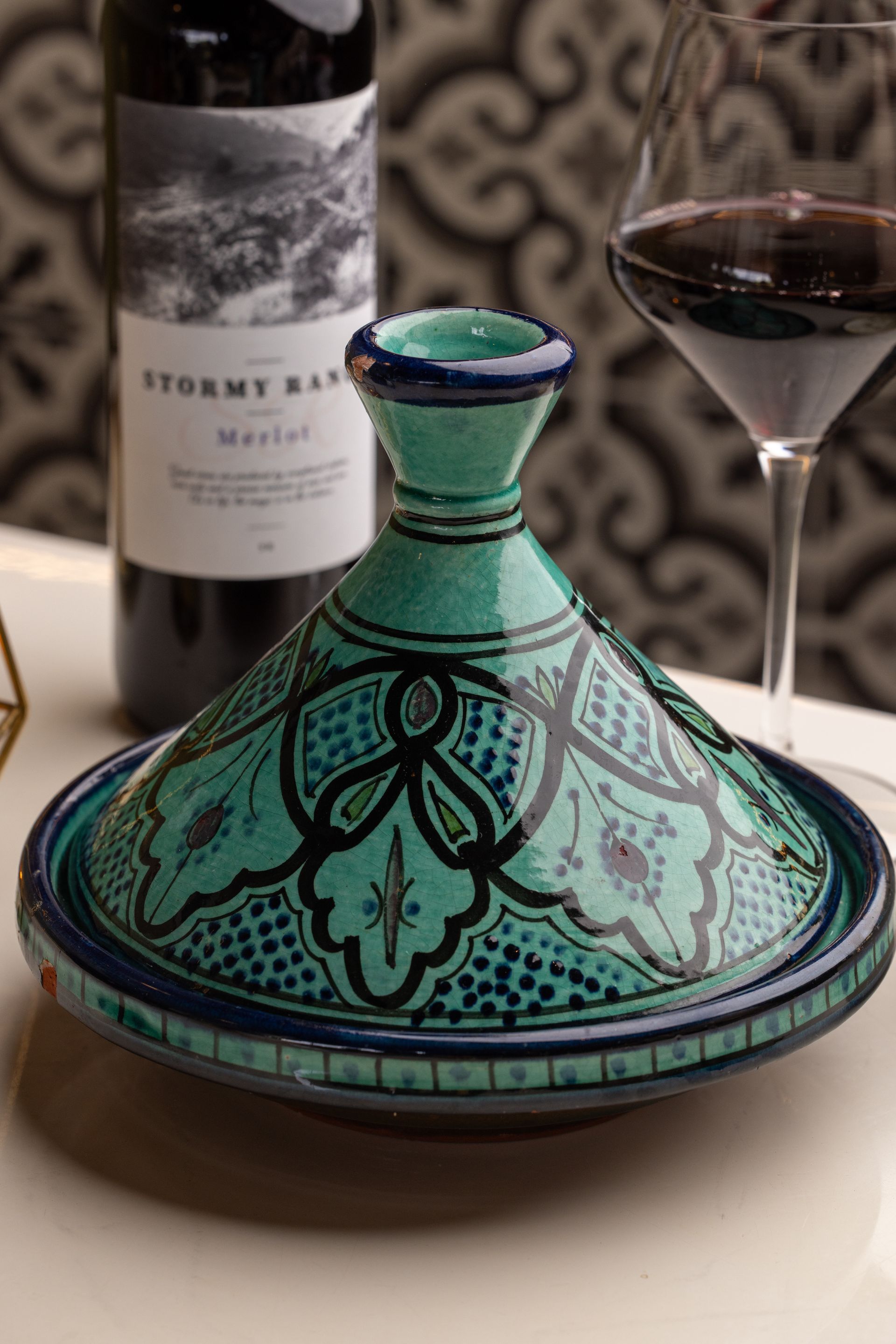 Green and blue Moroccan tagine pot with wine and bottle on a table.