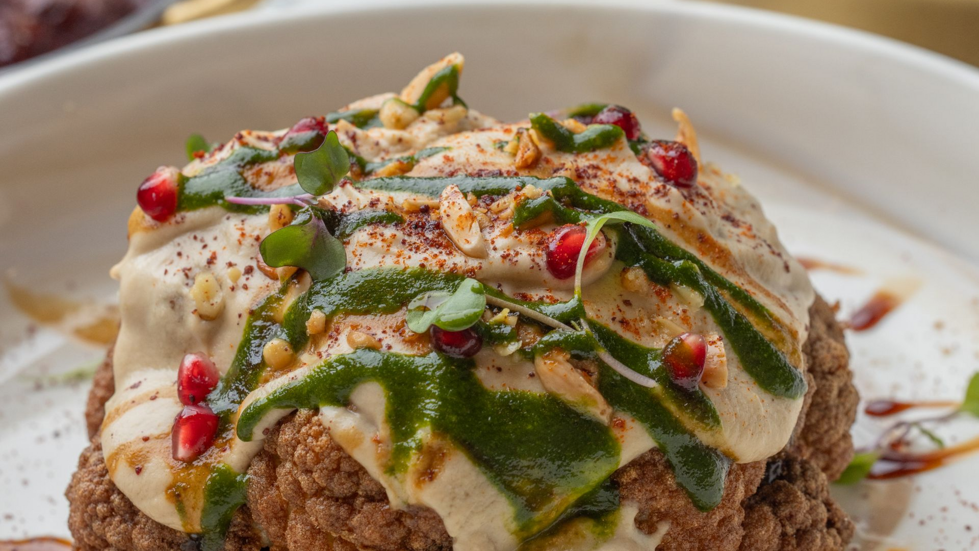 Close-up of a plate with a cauliflower dish, topped with sauces, pomegranate seeds, and nuts.