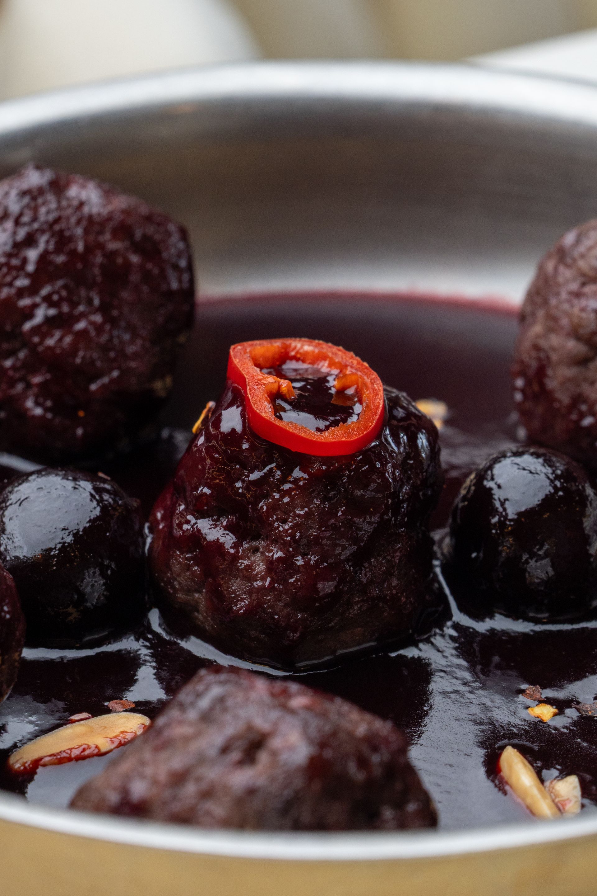 Meatballs in dark sauce, garnished with a red pepper slice, in a metallic bowl.