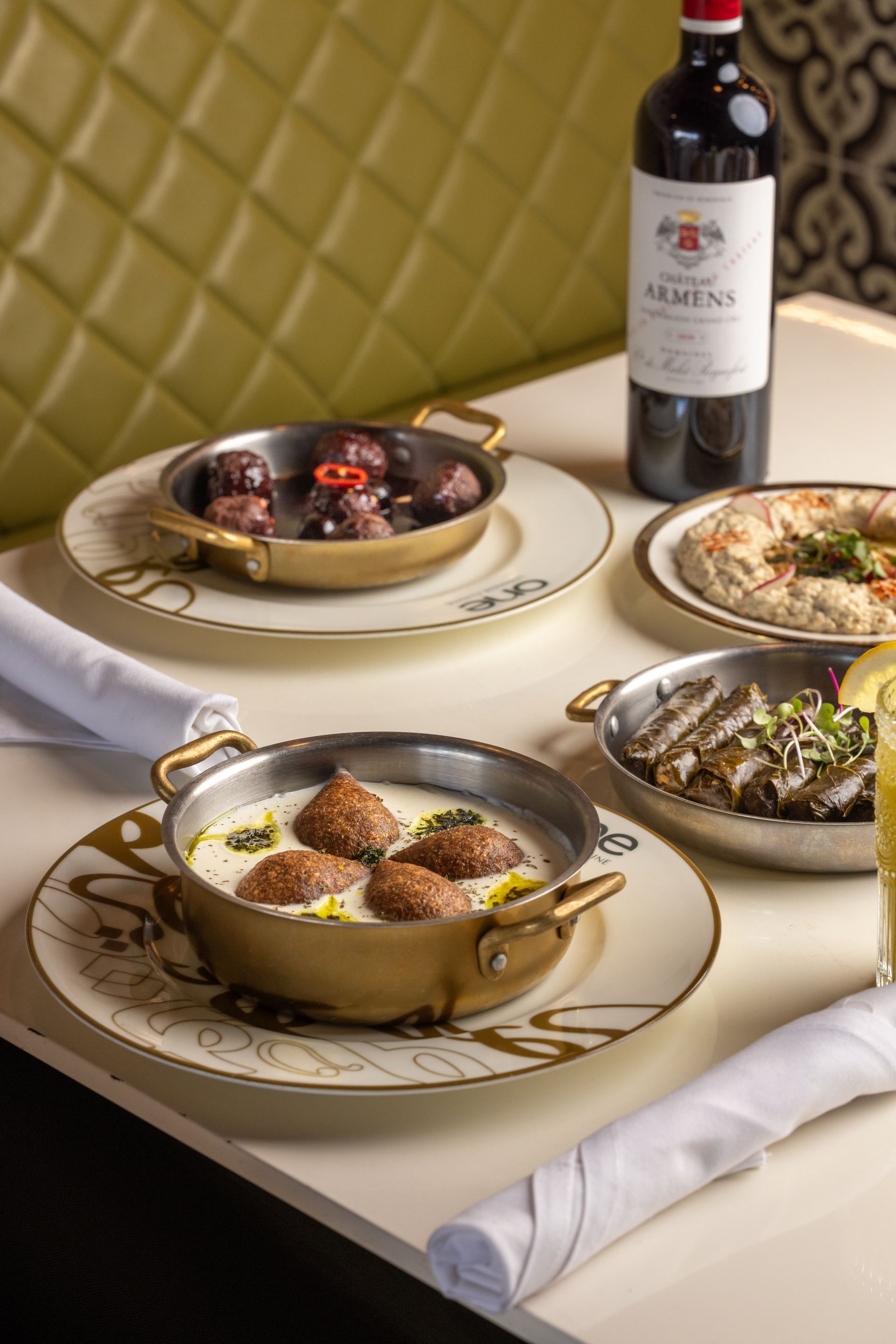 Several dishes in brass pans on a table with a wine bottle, and two rolled napkins.