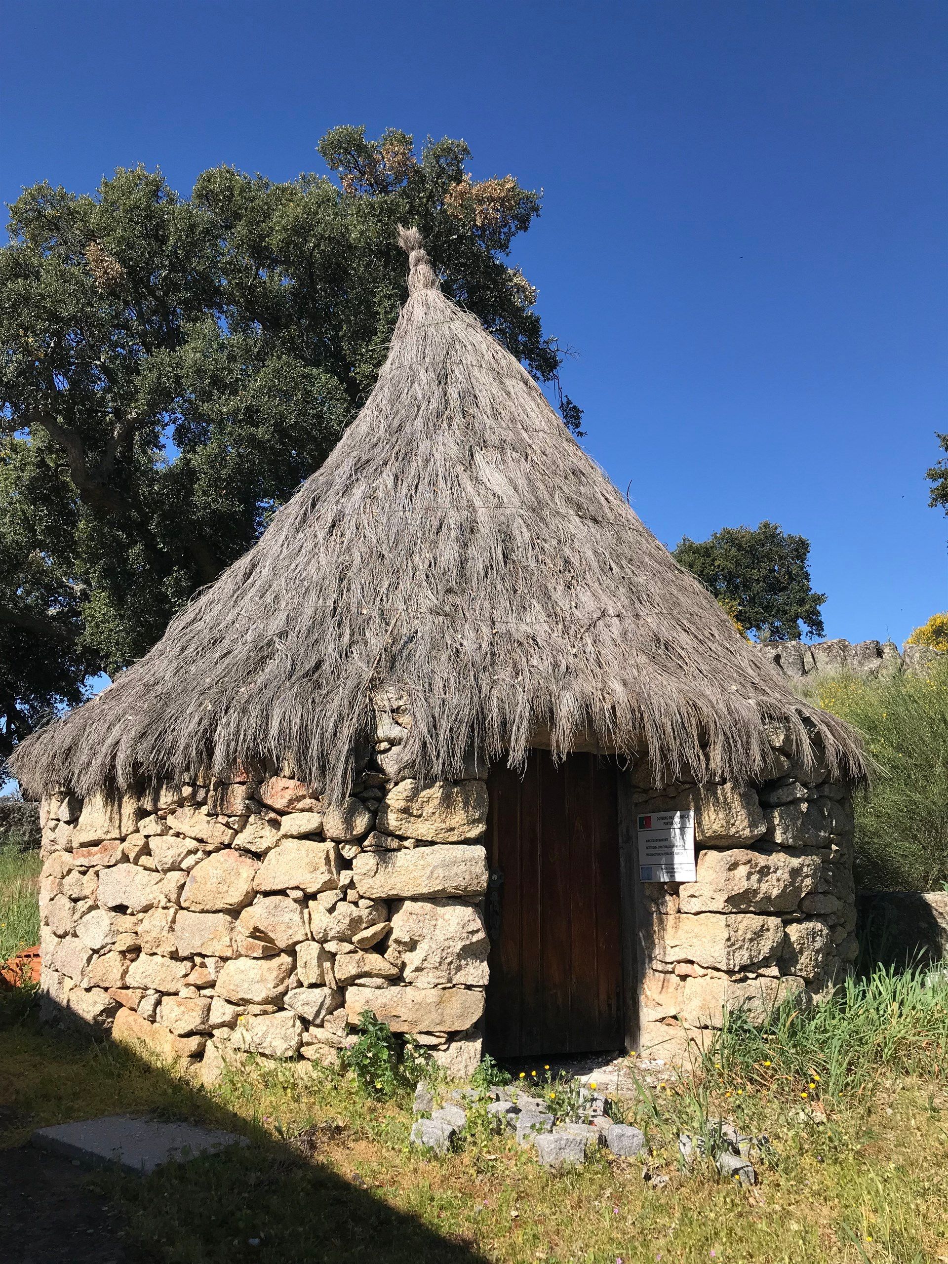 Uma cabana de pedra com telhado de palha e uma árvore ao fundo. Marvão 