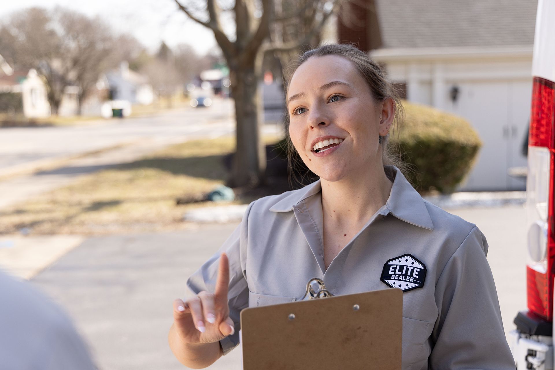 Woman in gray uniform holding clipboard, gesturing with finger near a van in a residential setting.