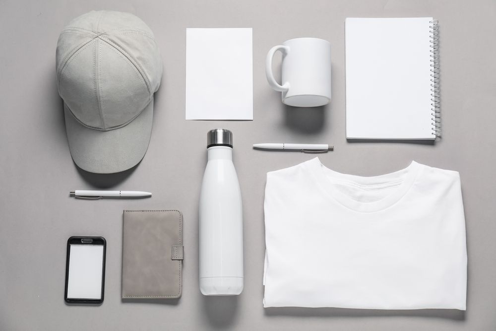 Flat lay of white branded merchandise: cap, notepad, mug, paper, notebook, pen, water bottle, phone, journal, and t-shirt.