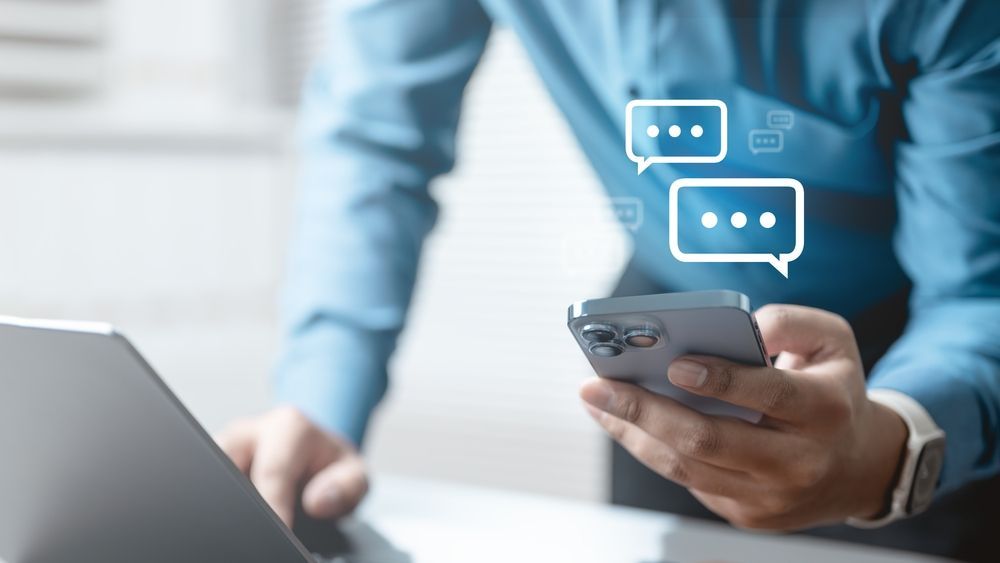 Person in blue shirt holds a phone, viewing messages. Laptop on the left.