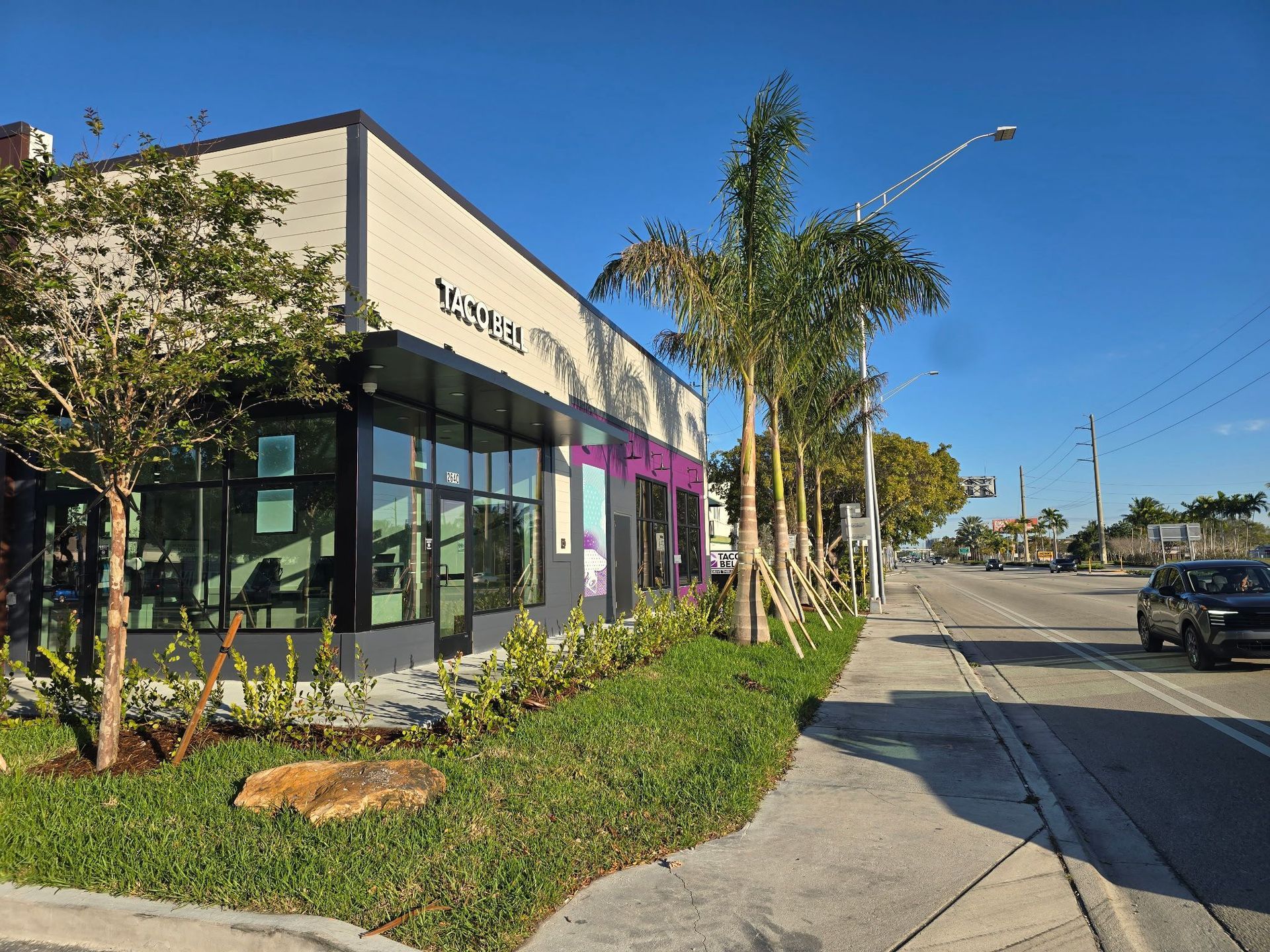 A modern building with large windows and a sign, next to a sidewalk and a road. Palm trees and other greenery are in front.