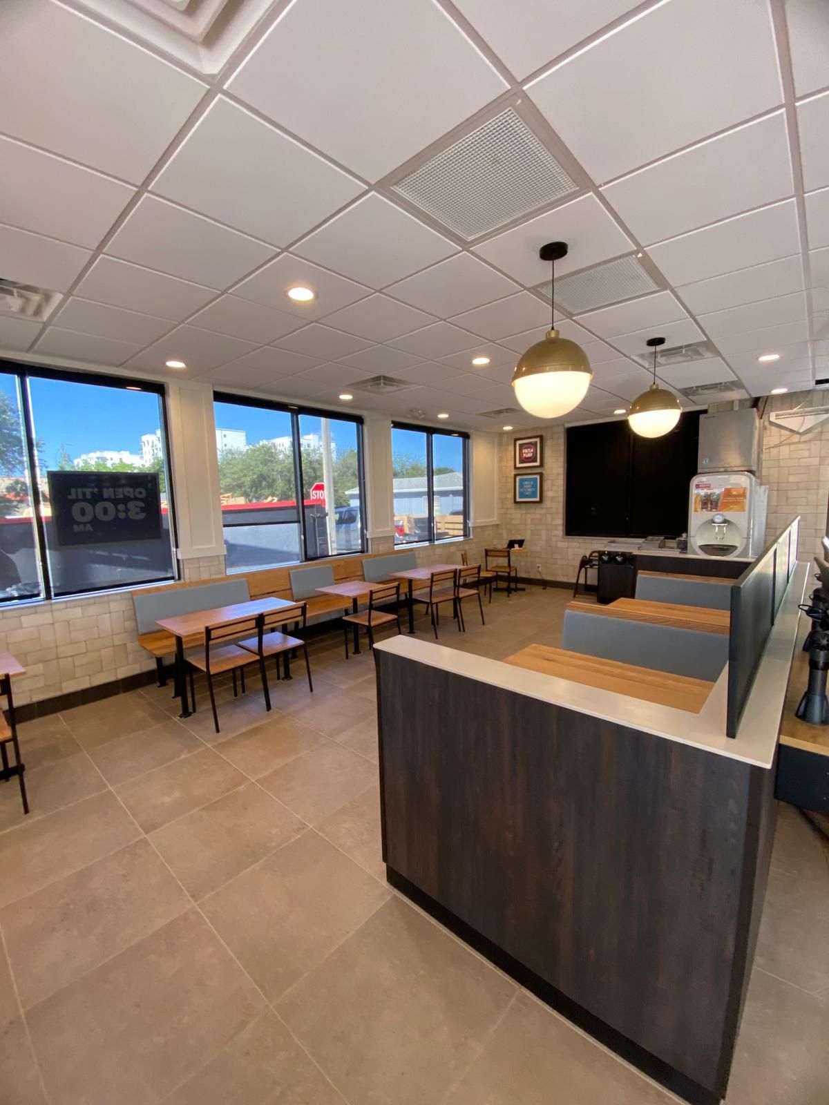 Empty, well-lit restaurant interior with tables, a counter, and large windows. Beige tile floor and white ceiling.