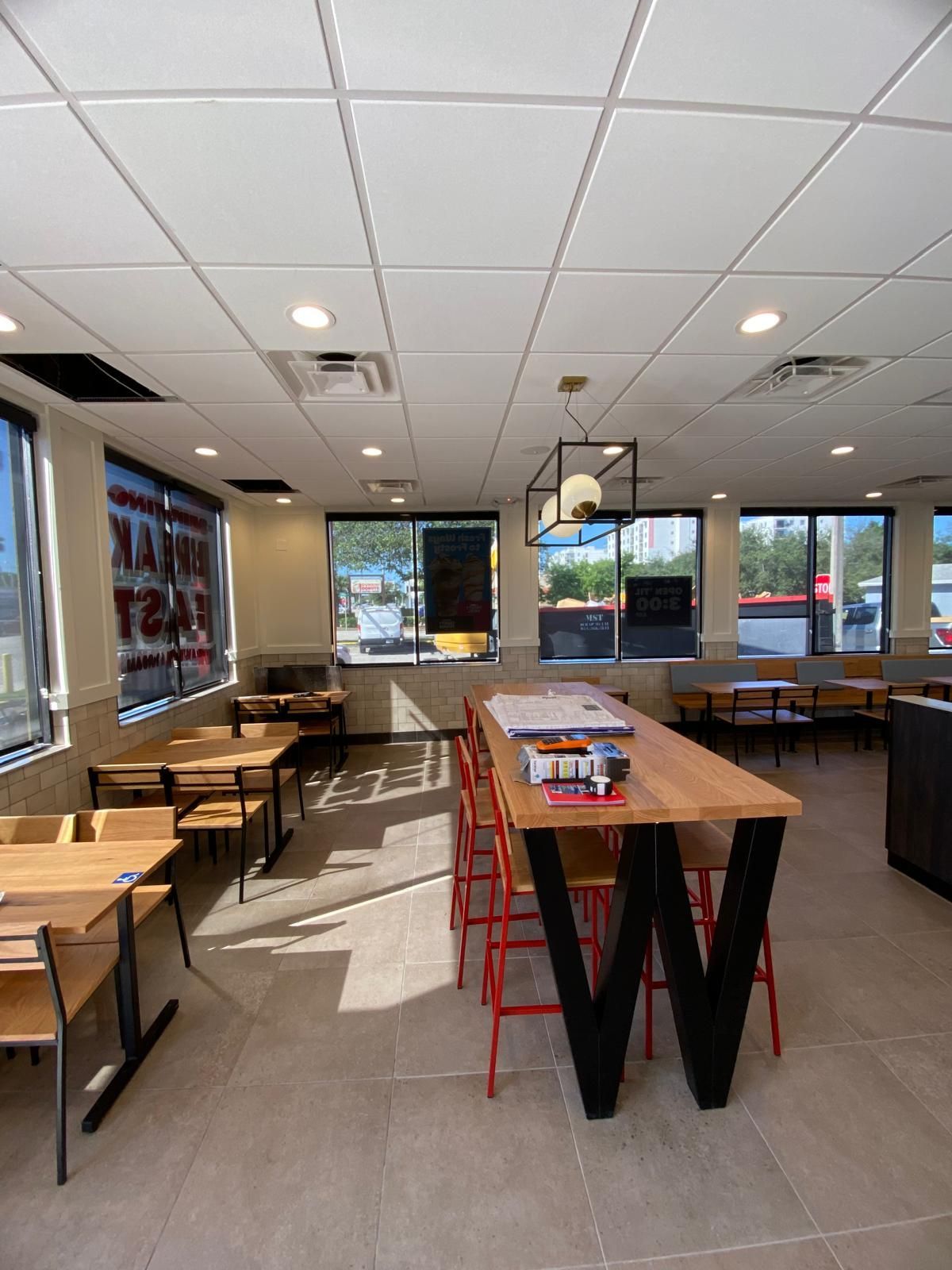 Interior of a restaurant with wooden tables, red chairs, and a long communal table under a light fixture.