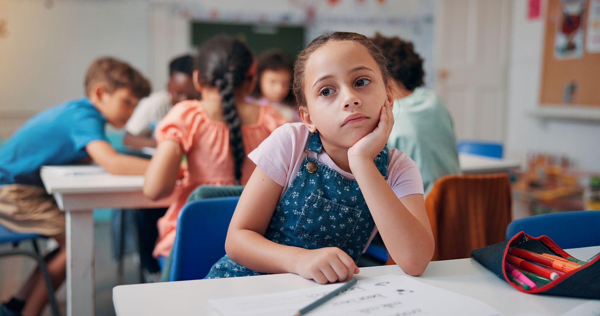 Bored student resting her head on her hand at a classroom desk with worksheet and pencil case.
