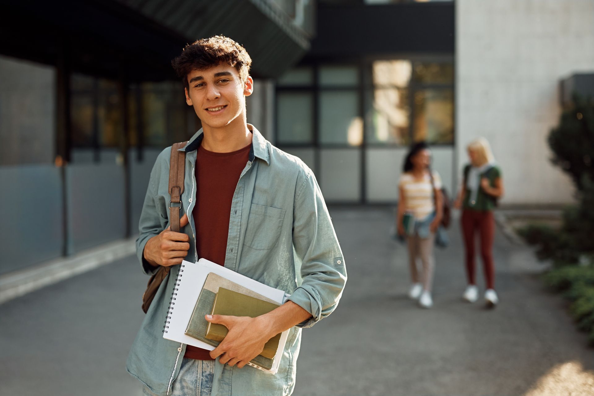 A student stands in the foreground holding books and a backpack, with two other people walking in the blurred background.