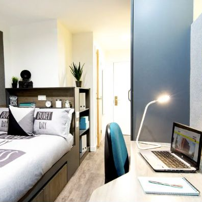 A bedroom with a bed , desk , laptop and lamp.