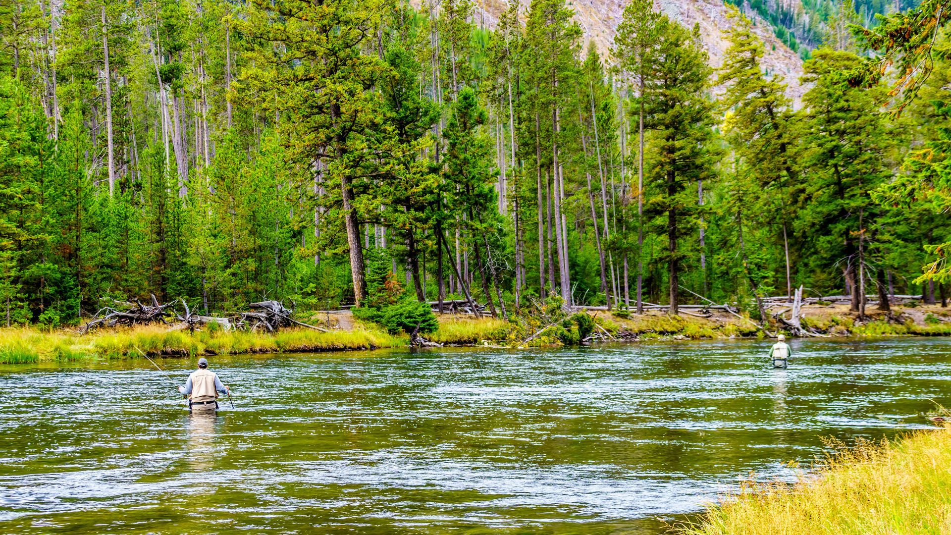 Two people are fishing in a river with mountains in the background.