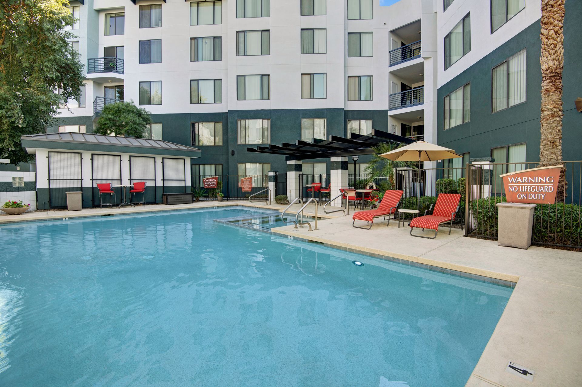 A large swimming pool surrounded by chairs and umbrellas in front of a building.