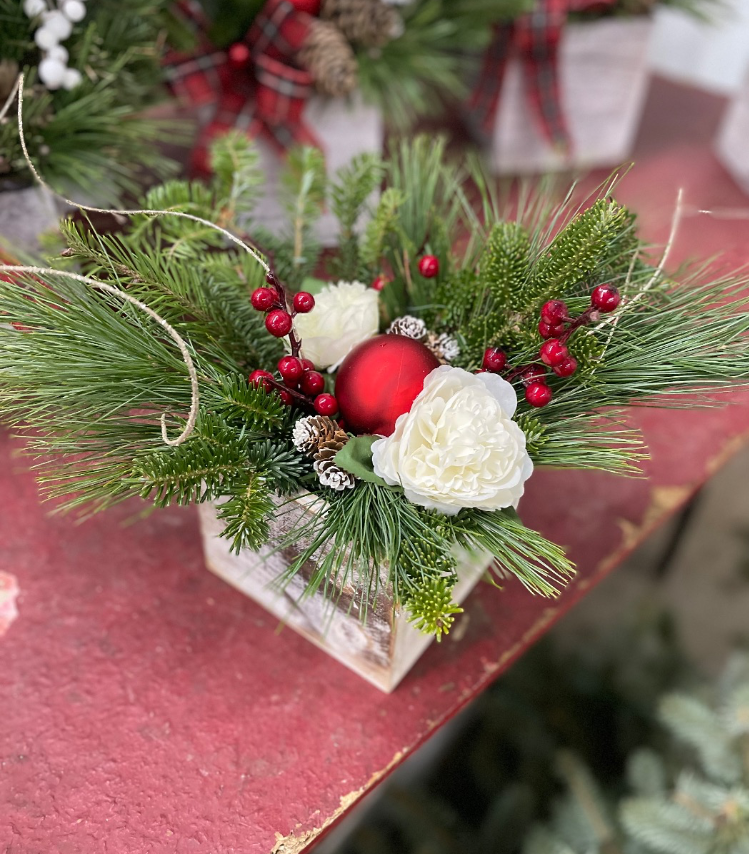 A holiday floral arrangement in a wooden box with pine branches, white flowers, red berries, and a shiny red ornament.