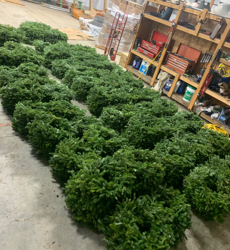 Numerous green, round evergreen wreaths are arranged in rows on a concrete floor inside a workshop or warehouse.