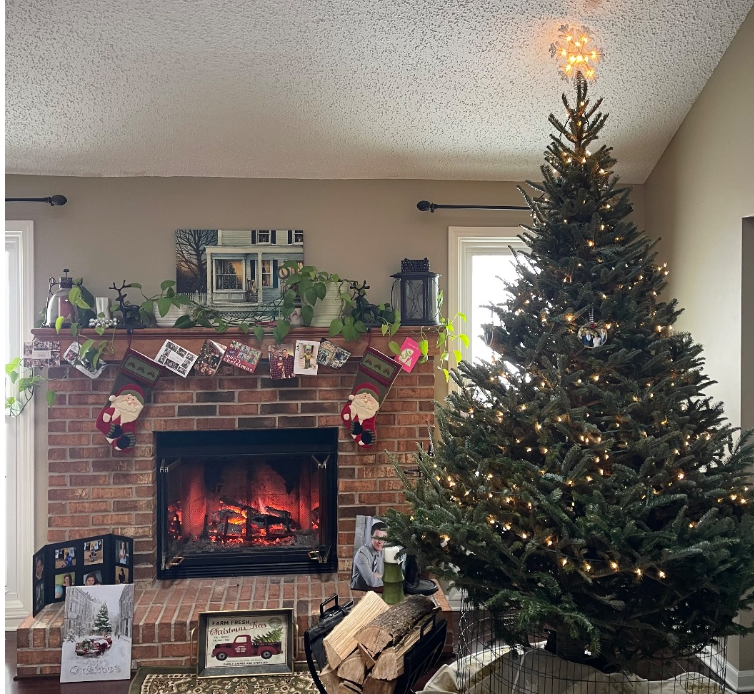 A lit Christmas tree next to a brick fireplace decorated with stockings, greenery, photos, and a framed holiday picture.