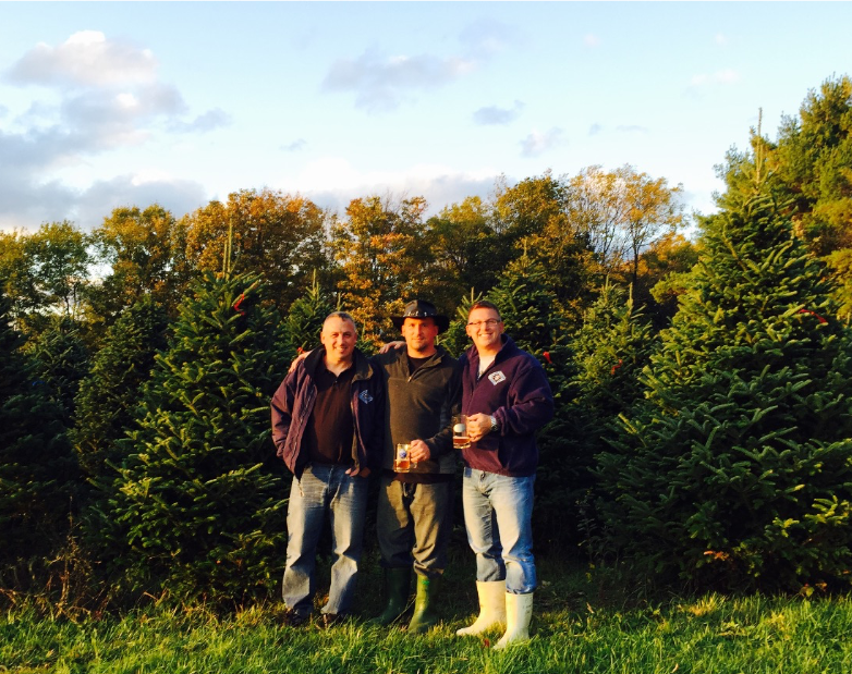 Three people stand side-by-side in a lush green Christmas tree farm during late afternoon.