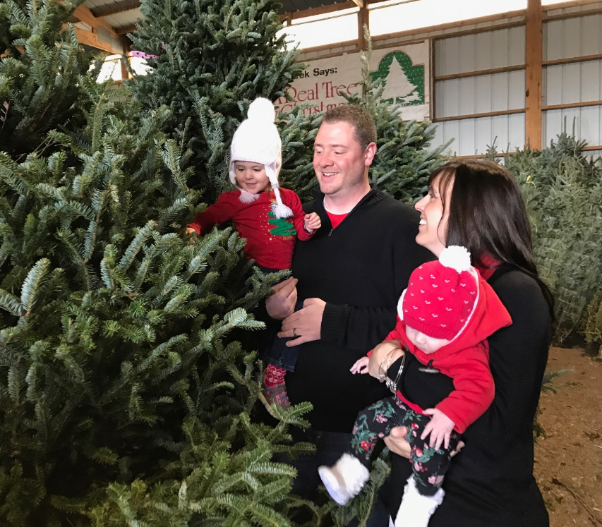 A parent holds two children in winter hats and sweaters while selecting a Christmas tree in an indoor lot.