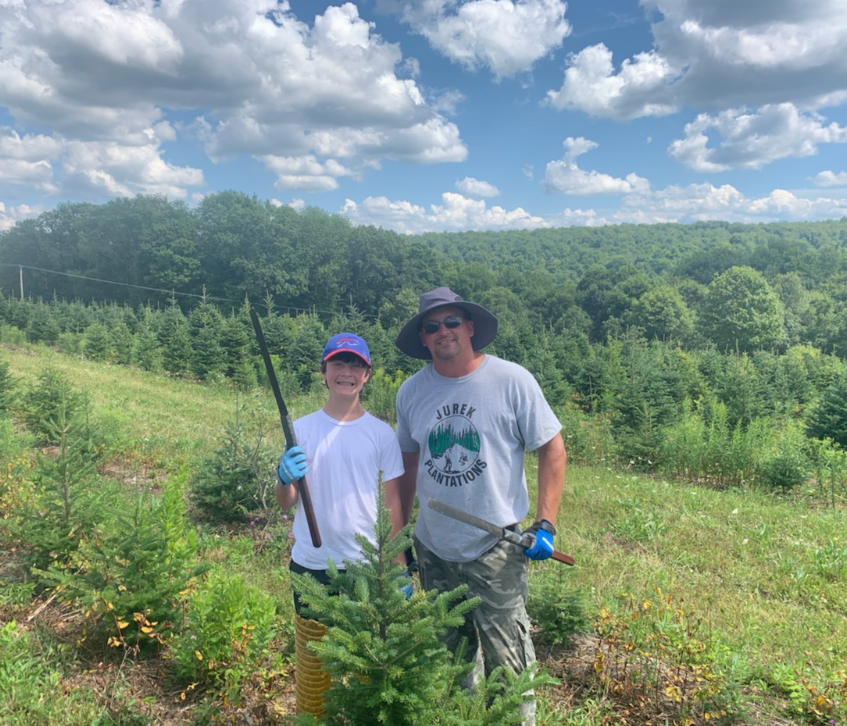 Two people planting trees in a field with a forest background under a cloudy sky.