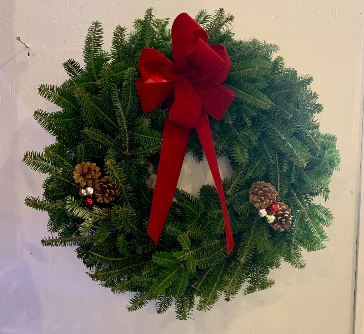 A circular evergreen wreath with a large red velvet bow and clusters of pinecones on a plain white door.