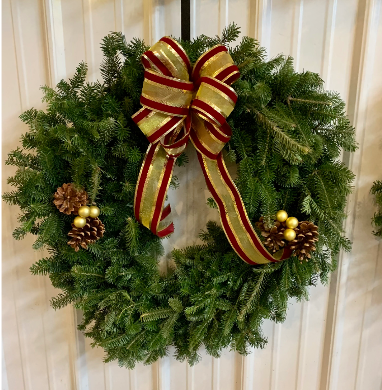 A holiday wreath made of evergreen needles, adorned with a large red and gold ribbon bow, pinecones, and gold ornaments.