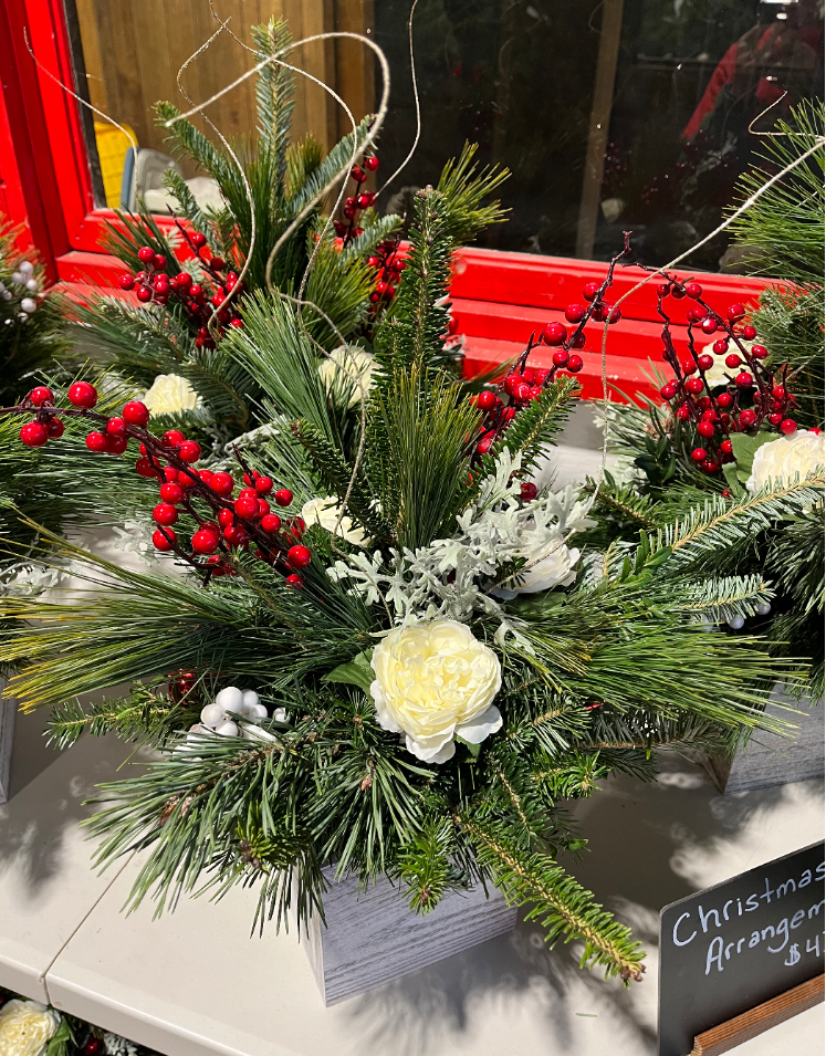 A festive holiday floral arrangement with white flowers, pine branches, and red berries in a white decorative box.