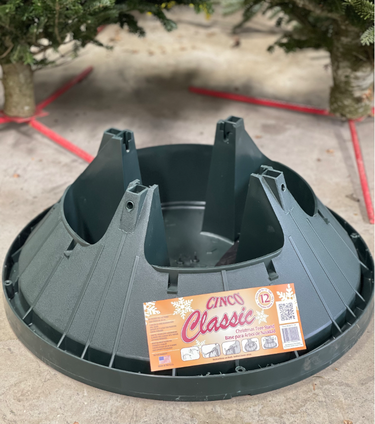 A dark green plastic Cinco Classic Christmas tree stand on a concrete surface, with a product label on the front.