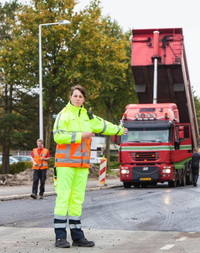 Een vrouw in een geel jasje staat voor een rode kiepwagen