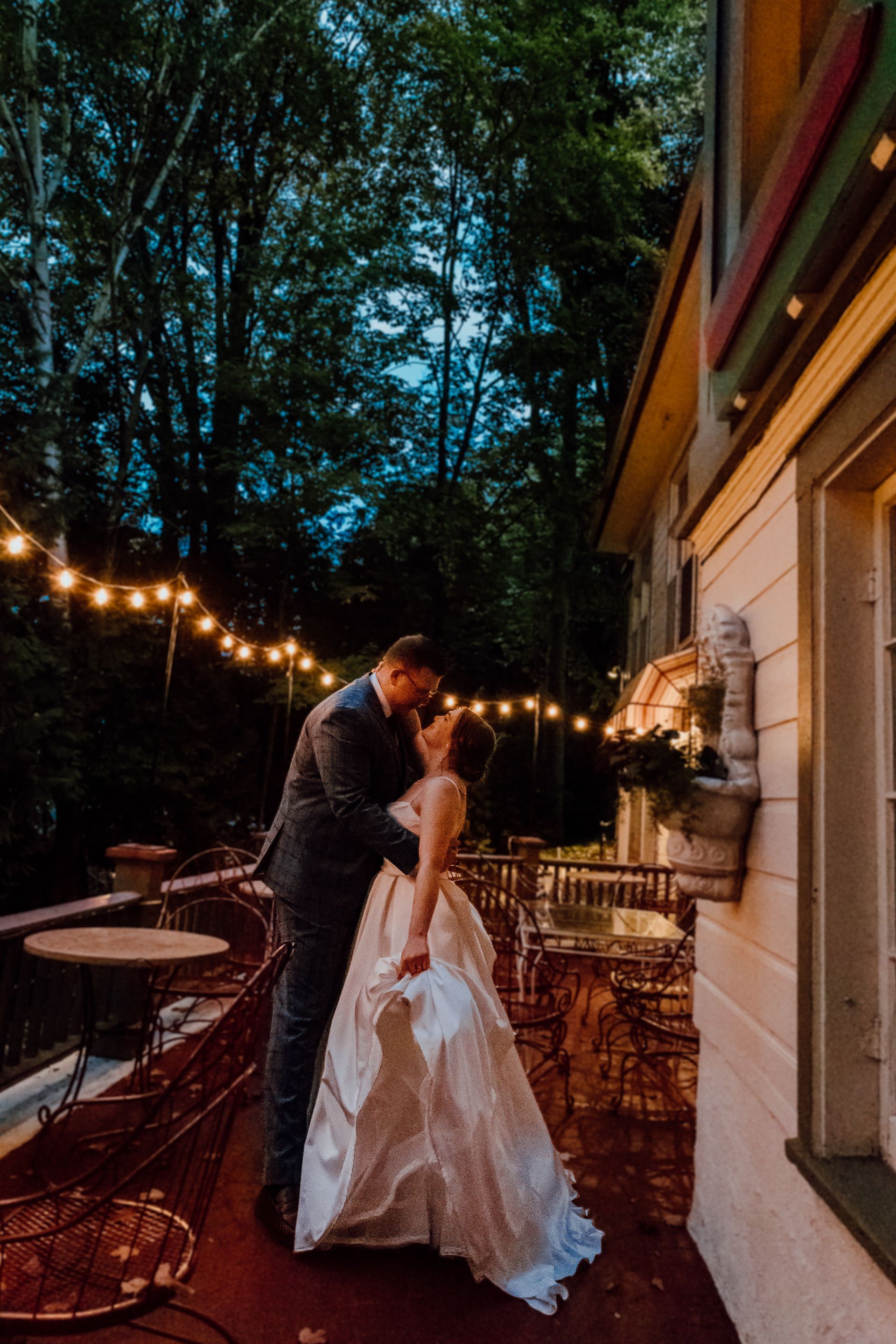 A bride and groom are kissing on a balcony under a string of lights.