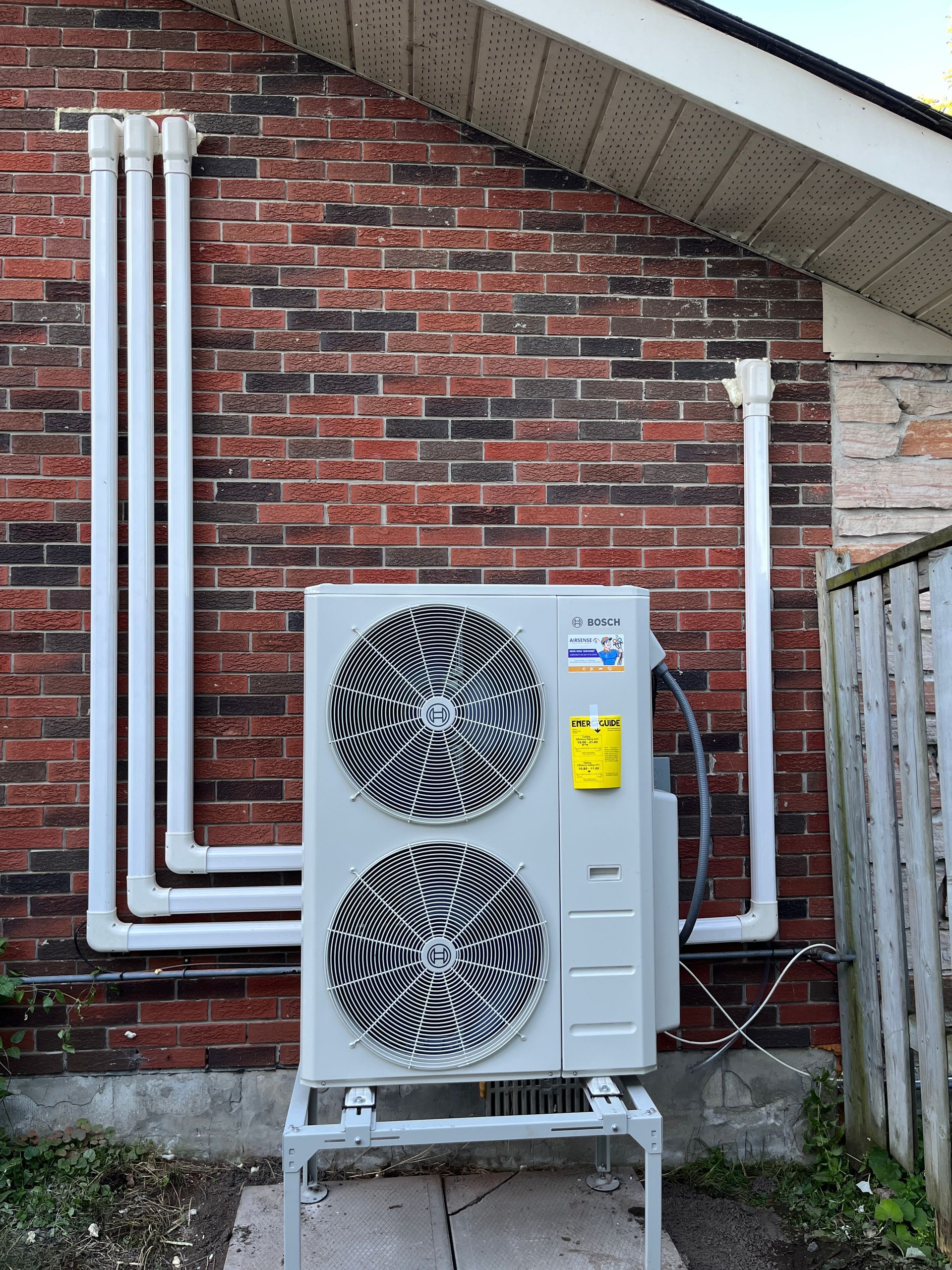 Heat pump unit mounted on a stand against a brick wall, white pipes connect to it.