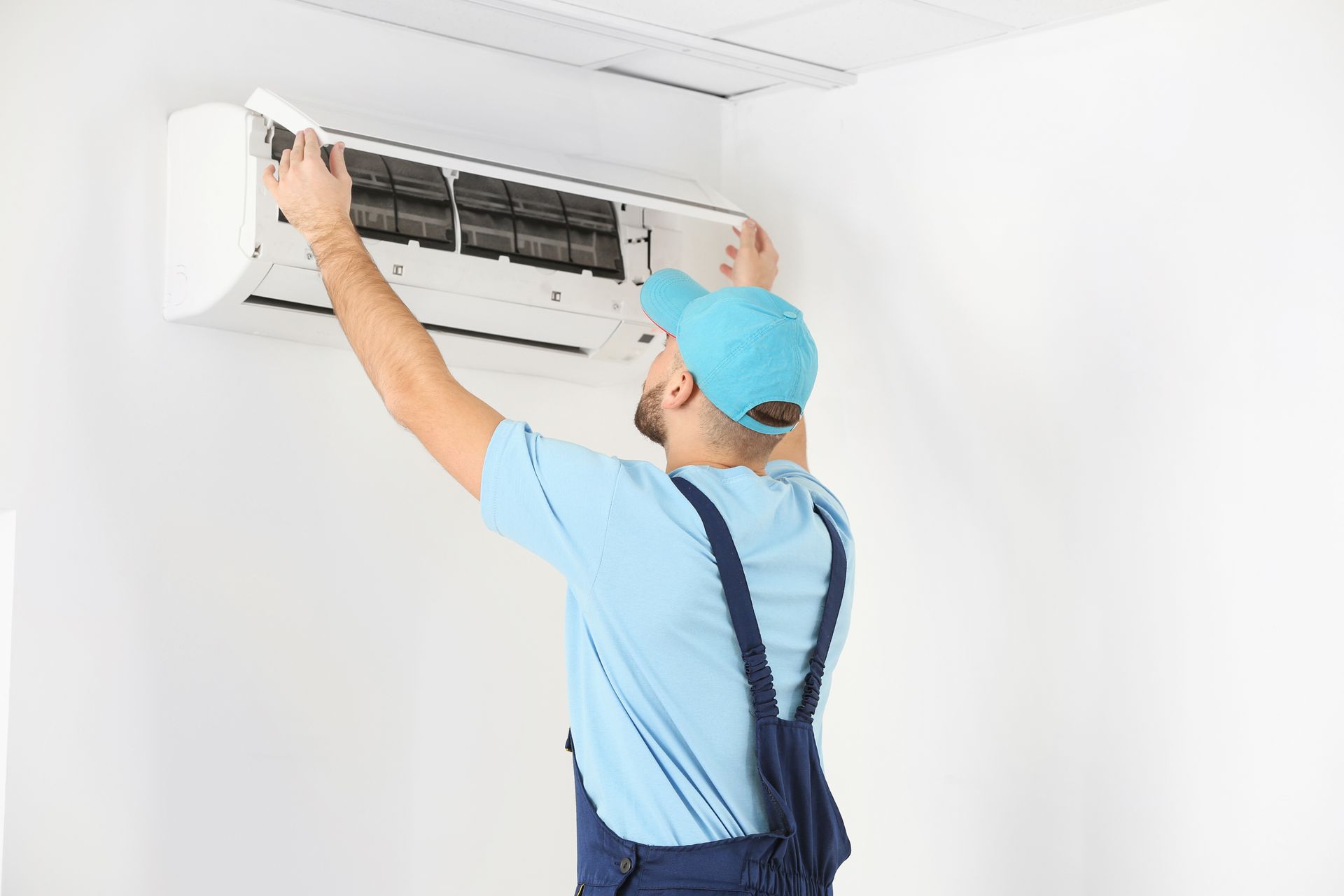 Man in blue overalls installing a white air conditioner on a white wall.